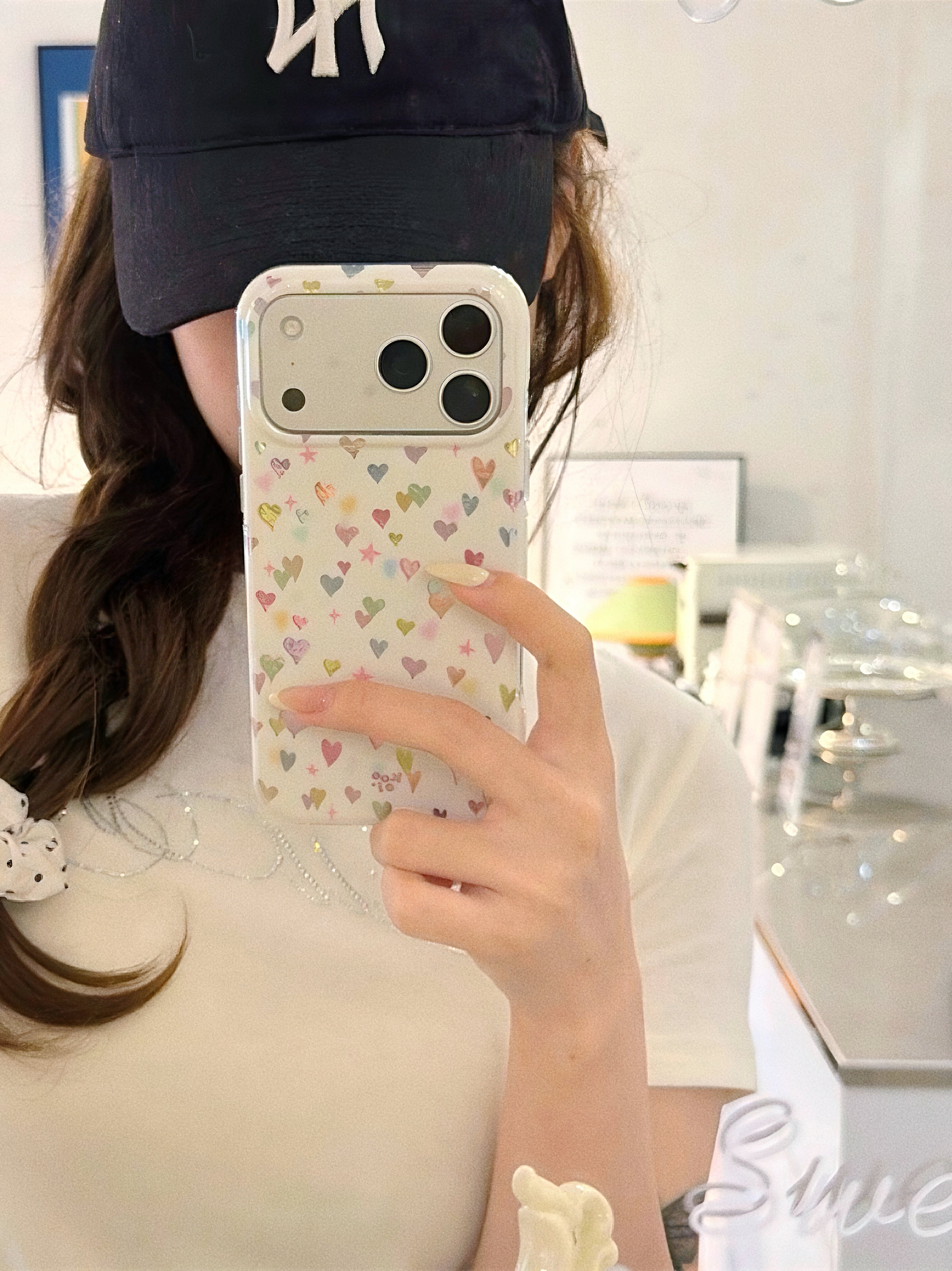 Mirror selfie showing a person holding a cream-colored hearts printed phone case for iPhone 17 Pro.